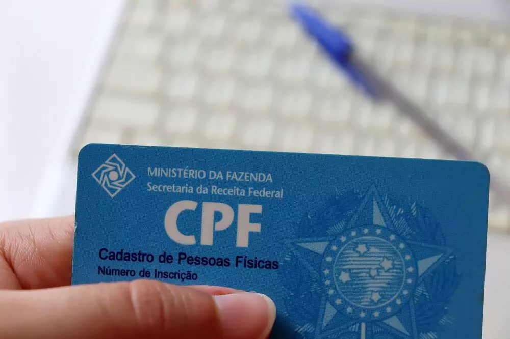 Mão segurando CPF com teclado e caneta ao fundo